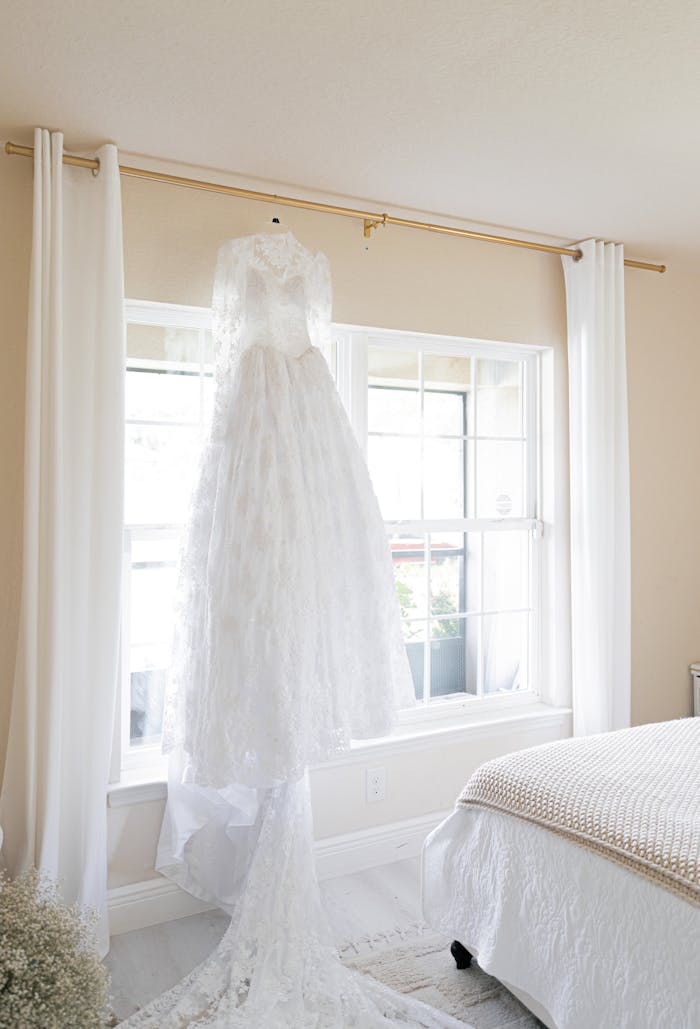 A luxurious white wedding dress hangs in a stylish, sunlit bedroom interior.